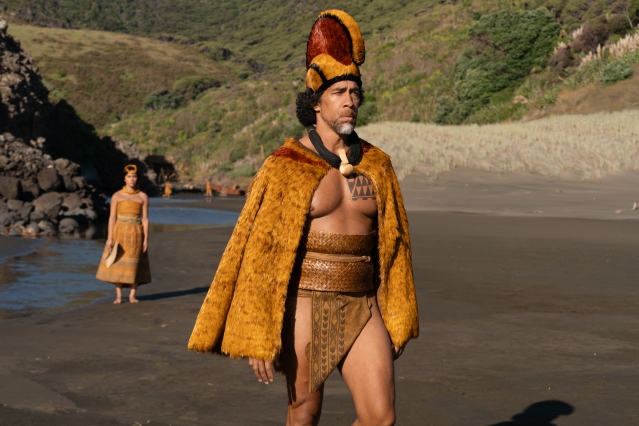 A Māori warrior in traditional battle attire walks across a black sand beach, with another figure visible in the background.